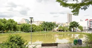 Vista de um parque em Nova Serrana, com um lago sereno no centro e bancos dispostos ao redor. Ao fundo, alguns prédios.