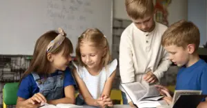 Quatro crianças brancas pequenas lendo livros em sala de aula. Ao fundo, um quadro branco com escritos.