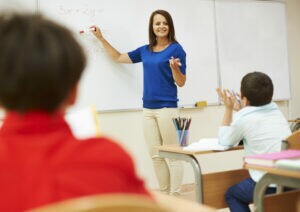 Professora explicando equação matemática no quadro enquanto alunos participam da aula. - Concurso SEE PB