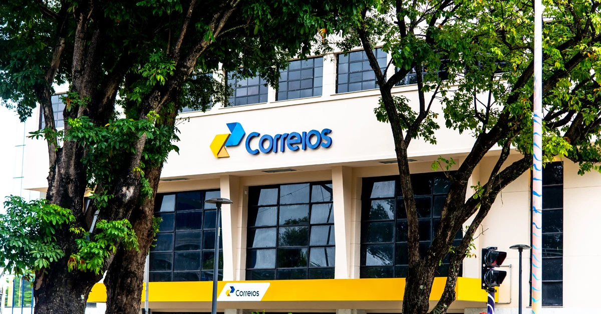 Fachada de um prédio dos Correios, com a logo da instituição e árvores em frente