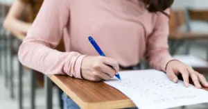 Mulher jovem concentrada escreve em uma folha de papel enquanto está sentada à mesa, em um ambiente de sala de aula.