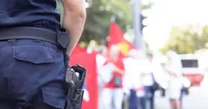 Agente da polícia em uniforme, observando uma multidão, enquanto manifestantes aparecem desfocados ao fundo.