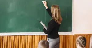 Professora escrevendo com giz em um quadro negro, explicando um tema para uma turma de alunos.