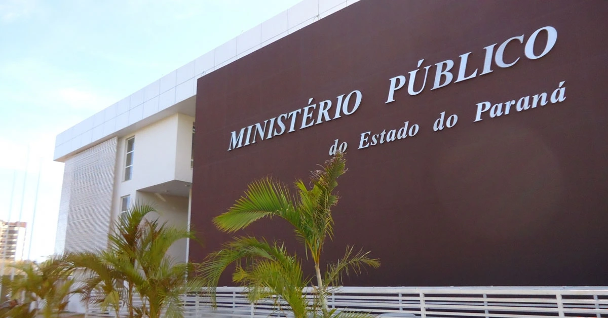 Fachada marrom com placa metálica escrito Ministério Público do Estado do Paraná. Cercado por grade branca e folhagem verde