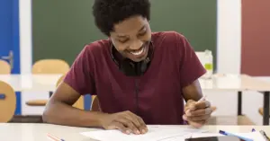 Homem negro jovem e sorridente escrevendo em um papel, em uma sala de aula.