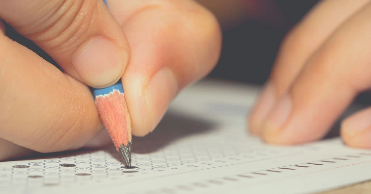 Estudante escrevendo com lápis em um papel durante uma prova na sala de aula / Concurso MPU