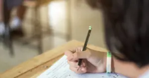 Estudante escrevendo com lápis em um papel durante uma prova na sala de aula / Concurso Prefeitura de Campos Altos