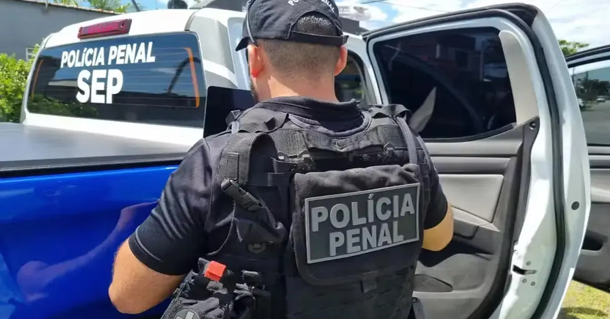 Um policial penal em um uniforme na caro preto do órgão em frente a viatura / Concurso Polícia Penal RJ