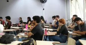 Sala de aula com vários estudantes sentados em carteiras individuais, prestando atenção e fazendo anotações / CNU 2025