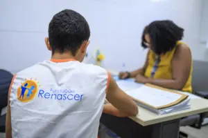 Atendimento na Fundação Renascer, com jovem de costas e funcionária preenchendo documentos. - Concurso Fundação Renascer