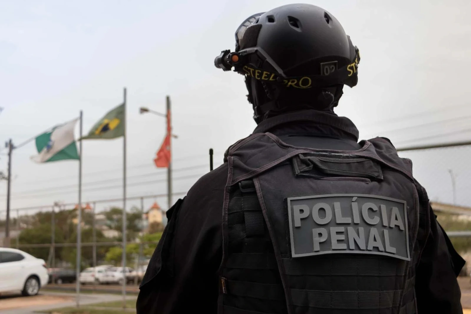 Aprovado no concurso Policia Penal PB fardado, de costas e olhando para três bandeiras.